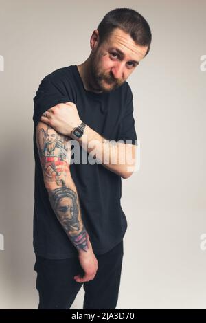 Il millenario europeo in abiti neri mostra il suo braccio tatuato al cowboy fotocamera girato sfondo grigio studio scatto . Foto di alta qualità Foto Stock