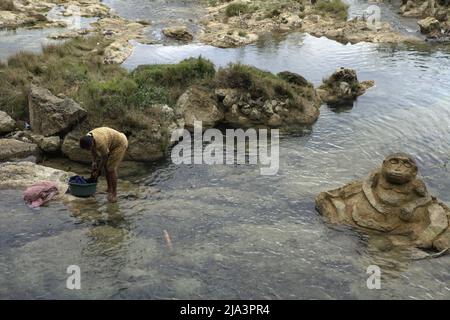 Una donna che lava abiti su un ruscello vicino a una pietra scolpita di figura umana vicino a Waikelo Sawah, una fonte d'acqua rara a Sumba, un'isola regolarmente colpita dalla siccità, che si trova nel villaggio di Tema Tana, East Wewewa, Southwest Sumba, East Nusa Tenggara, Indonesia. Foto Stock