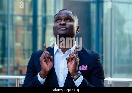 Bell'uomo d'affari Afro in un elegante abito nero che guarda nel cielo e prega sullo sfondo della strada grattacielo ufficio. Fortuna e successo affare Foto Stock