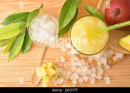 Particolare di un bicchiere con succo di mango con ghiaccio su un tavolo di legno con frutta e ghiaccio tritato intorno ad esso. Vista dall'alto. Composizione orizzontale. Foto Stock