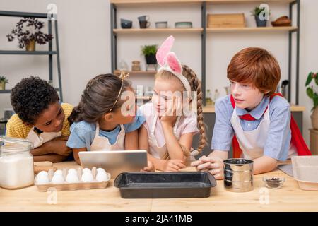 I bambini si sono avvolti nella ricetta di lettura con l'aiuto di un tablet digitale mentre si trovano in piedi al tavolo da cucina in legno per fare sorpresa per il Mothers Day Foto Stock