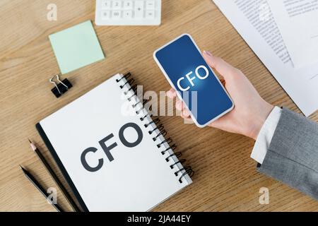 vista dall'alto di una donna che tiene uno smartphone con scritta cfo sullo schermo vicino a notebook e cancelleria Foto Stock