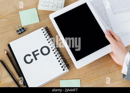 vista dall'alto di una donna che tiene un tablet digitale con schermo vuoto vicino al notebook con scritte e cancelleria cfo Foto Stock