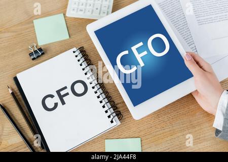 vista dall'alto di una donna che tiene un tablet digitale con scritte cfo sullo schermo vicino a notebook e cancelleria Foto Stock