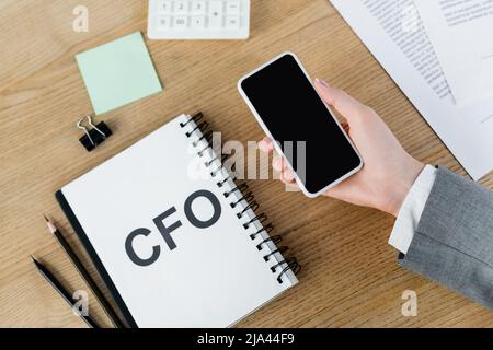 vista dall'alto di una donna che tiene uno smartphone con schermo vuoto vicino al notebook con scritte e cancelleria cfo Foto Stock