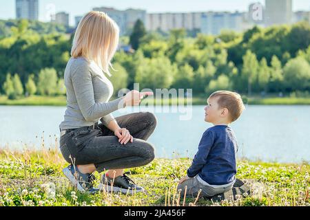 bella madre scarabei punti dito suo bambino carino (figlio) seduto sull'erba nel parco su uno sfondo di alberi verdi e fiume (lago) Foto Stock