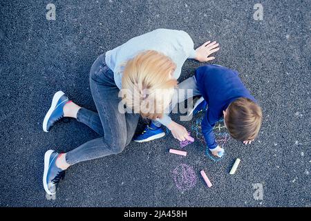 cute bambino figlio e madre dipinta con gesso colorato crayons sull'asfalto nel parco su uno sfondo sopra la vista Foto Stock