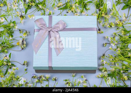 Confezione regalo grigio-blu con un arco su sfondo blu in una cornice di fiori selvatici Foto Stock