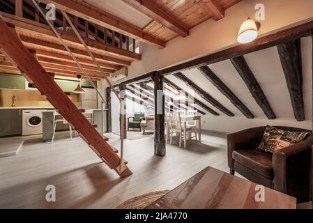 soggiorno di un appartamento monolocale con travi in legno e colonne sui soffitti inclinati, scala in legno e cucina aperta sullo sfondo Foto Stock