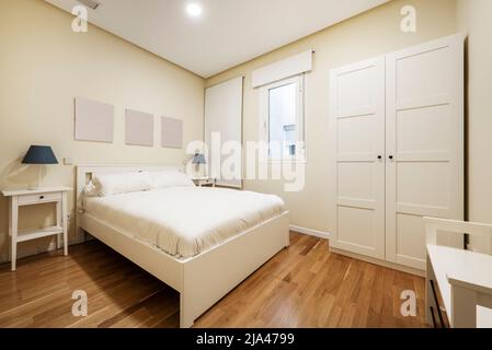 Camera da letto con letto matrimoniale in legno bianco, armadio a due porte abbinato, finestre con persiane e pavimento in parquet di quercia e piumino bianco Foto Stock