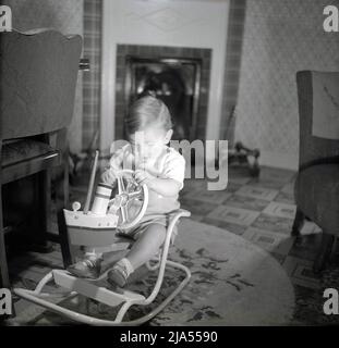 1950s, storico, in una sala d'ingresso, un bambino che gioca su una barca a tema giocattolo metallo incorniciato sedia a dondolo, Inghilterra, Regno Unito. Foto Stock