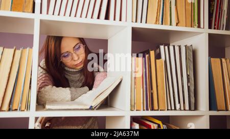Uno studente sta leggendo un libro nella biblioteca dietro le librerie. Una giovane donna premurosa in piedi in una biblioteca tra i libri e la lettura di un libro di testo A. Foto Stock