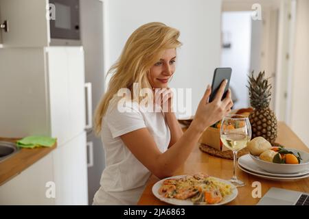 Ritratto della giovane bionda caucasica che tiene smartphone in cucina durante la cena. Foto Stock