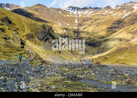 Un sentiero stretto qui conduce attraverso una valle stretta, fiancheggiata da pendii innevati e una cascata lontana, verso la piscina Seljavallalaug ai piedi del vulcano Eyjafjallajökull; un escursionista con una giacca verde si avvicina attraverso un campo di massi. Costruita nel 1923 dal contadino Björn Andrésson, la piscina di 25 per 10 metri è considerata una delle più antiche piscine sopravvissute in Islanda ed era la più grande del paese fino al 1936. Foto Stock