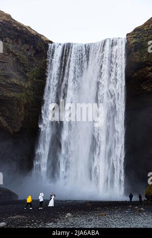 La cascata di Skógafoss alta 60 metri è mostrata qui in tutto il suo splendore, che si erge sopra una sessione fotografica di nozze; la sposa e lo sposo appaiono minuscoli di fronte alla cascata, con due fotografi in giallo e bianco in primo piano a sinistra e due visitatori ai bordi dell’inquadratura a destra. Secondo la leggenda, il colono vichingo Þrasi Þórólfsson seppellì una cassa del tesoro qui in una grotta dietro la cascata, da cui è stato recuperato solo un anello – ora conservato nel museo di Skógar. Foto Stock