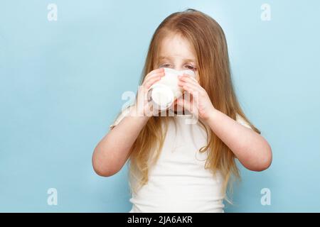 piccola bella ragazza che beve latte in un bicchiere su sfondo blu Foto Stock