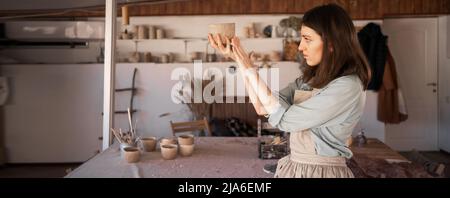 giovane artigiano ceramista si è concentrato sulla scultura di una tazza di argilla cruda durante una lezione di ceramica in uno studio creativo. il proprietario di affari di ceramica femminile fa l'artigianato Foto Stock