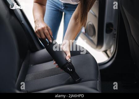 Donna che aspira il sedile posteriore nella sua auto nel garage di casa. Femmina che utilizza un aspirapolvere portatile per rimuovere polvere e sporcizia. Pulizia dell'abitacolo dell'auto Foto Stock