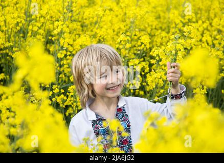 Carino gioioso ragazzo ucraino di 6 anni in una tradizionale camicia ricamata si erge tra un campo di colza giallo fiorito. Bambini per la Pace. Arresto t Foto Stock