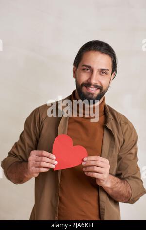 Verticale vita in alto ritratto di bel borsato giovane uomo che tiene rosso carta cuore sorridente alla fotocamera, copia spazio Foto Stock