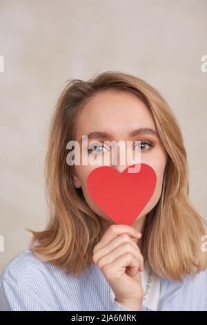Primo piano ritratto studio di affascinante giovane donna nascondendo la parte inferiore del suo viso con cuore di carta guardando la fotocamera, spazio copia Foto Stock