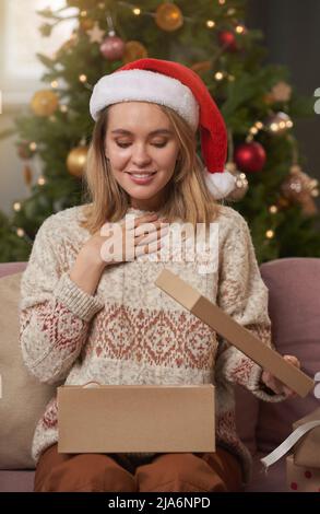 Verticale medio ritratto di felice donna caucasica indossando casual caldo maglione e rosso Santa cappello seduto sul divano godendo unboxing fantastici regali per CH Foto Stock