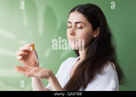 Una giovane bella donna applica olio cosmetico da una pipetta al palmo della mano. Sfondo verde con un'ombra da una foglia monstera. Il concetto Foto Stock