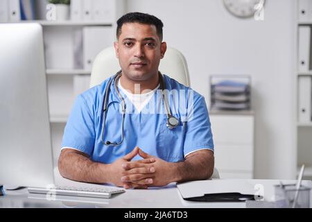 Ritratto orizzontale medio del bel giovane adulto medio orientale medico seduto alla scrivania nel suo ufficio guardando la macchina fotografica Foto Stock
