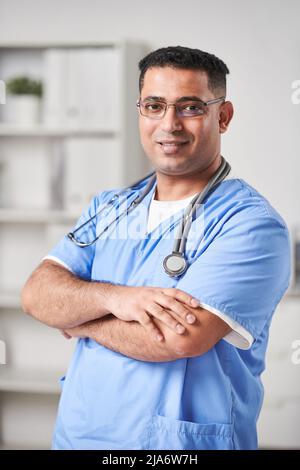 Verticale vita su ritratto scatto di giovane adulto moderno medio-orientale medico generico indossando uniforme blu e occhiali in piedi con le braccia croce Foto Stock
