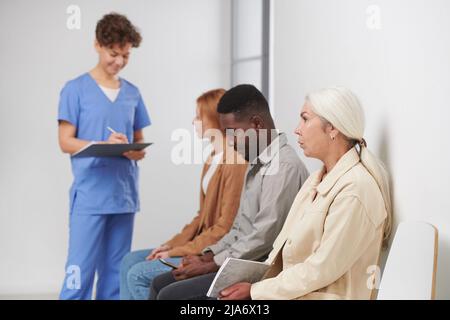 Gruppo di tre persone sedute in coda in attesa di consultazione in ospedale moderno, operatore medico in piedi vicino a loro facendo appunti Foto Stock