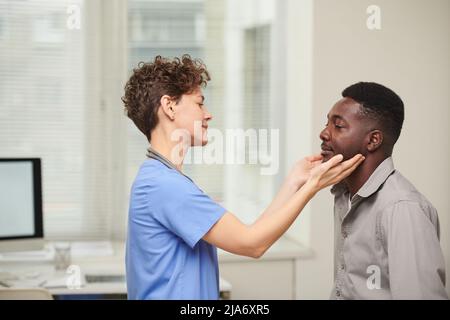 Vista laterale orizzontale shot di medico femminile indossando blu uniforme controllo dimensioni di pazienti afroamericani linfa ghiandole Foto Stock