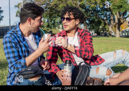 latino caucasico maschio omosessuale argentino coppia seduta sul prato nel parco sorridendo, godendo di bere compagno in una giornata di sole. Foto Stock