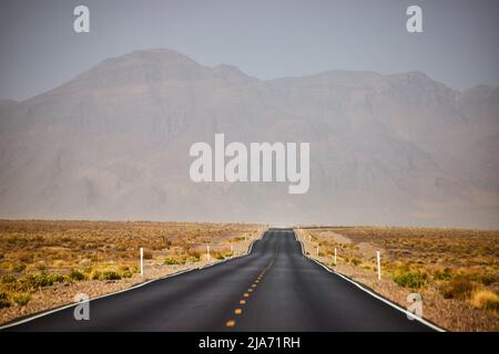 Strada lastricata nera che conduce attraverso il paesaggio sabbioso del deserto e le montagne Foto Stock