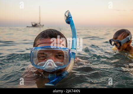 Ritratto di un giovane che nuota in mare dal suo yacht al tramonto snorkeling con suo figlio piccolo che indossa anche una maschera da nuoto. Foto Stock
