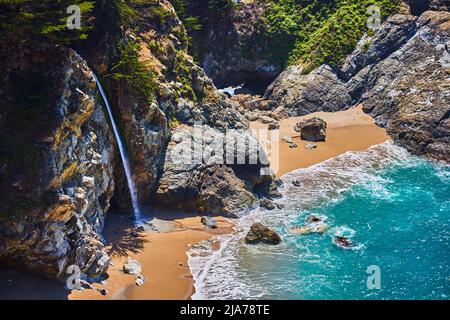 Rocce costiere con cascata che si stende sulle spiagge sabbiose accanto alla baia dell'oceano Foto Stock