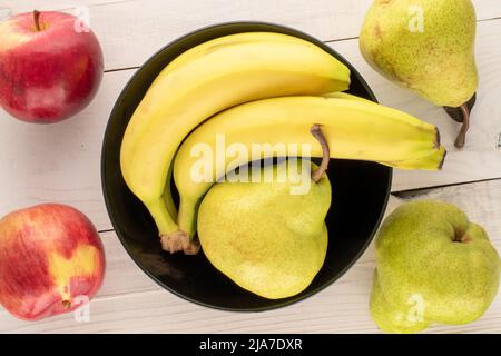 Tre succose pere biologiche, un mazzo di banane e due mele rosse con un piatto di ceramica nera su un tavolo di legno, primo piano, vista dall'alto. Foto Stock