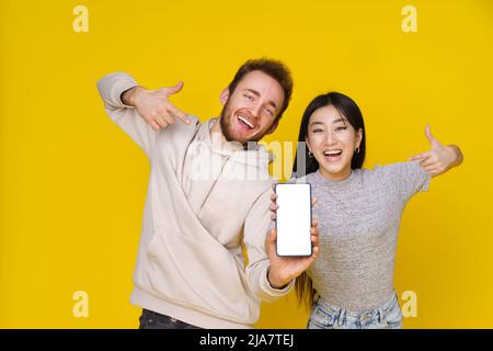 Ragazza asiatica e ragazzo caucasico che mostrano pollici in mano smartphone mostrando bianco schermo vuoto, app pubblicitarie mobile e sorriso eccitato sulla fotocamera isolato su sfondo giallo. Posizionamento del prodotto. Foto Stock