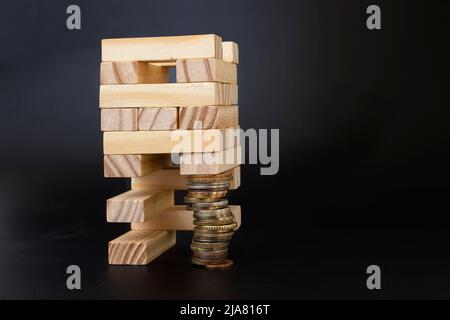 struttura di blocchi di legno supportata da una pila di monete. Costruzione gioco metafore isolato su sfondo nero. Foto Stock