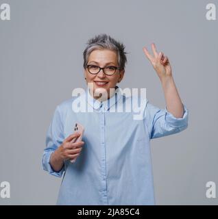 Donna d'affari matura dai capelli grigi con smartphone a portata di mano che guarda la fotocamera, che funziona, fa shopping online o controlla i social media. Bella donna in camicia blu isolato su bianco. Immagine a toni. Foto Stock