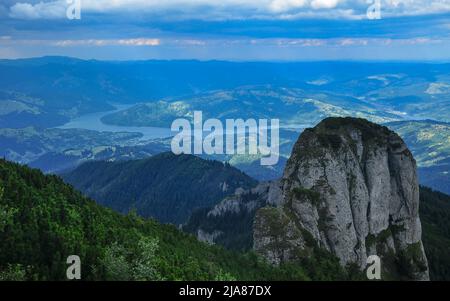 Una massiccia scogliera calcarea che sorge sopra le selvagge foreste di abete rosso dei Monti Ceahlau. Sole illumina le acque blu di un lago e le colline sottostanti Foto Stock