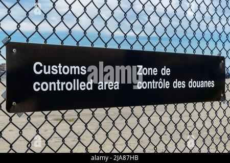 Un segno bilingue dell'area doganale controllata in inglese e francese su una catena di collegamento. Vista ad angolo con cielo blu sopra, parcheggio sotto. Foto Stock