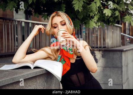 Giovane donna che indossa abito nero, fazzoletto di seta, in piedi alla ringhiera, sfogliando la rivista, bevendo una tazza di caffè. Foto Stock