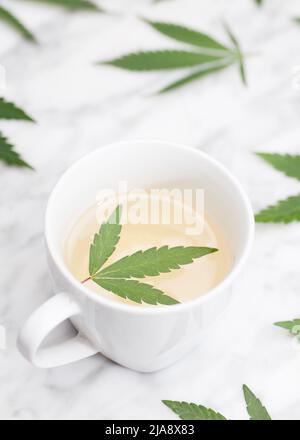 tazza bianca con infusione di marijuana con foglia verde all'interno su tavolo di marmo con foglie. vista dall'alto. Foto Stock
