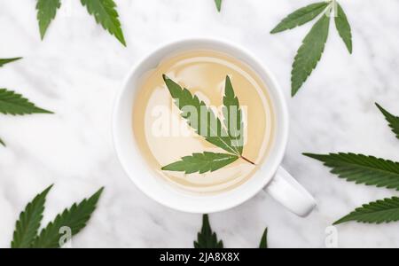 tazza bianca con infusione di marijuana con foglia verde all'interno su tavolo di marmo con foglie. vista dall'alto. Foto Stock