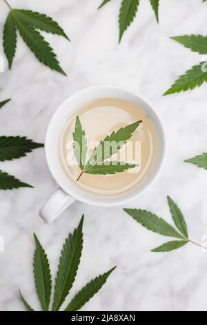 tazza bianca con infusione di marijuana con foglia verde all'interno su tavolo di marmo con foglie. vista dall'alto. Foto Stock