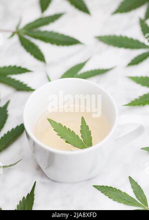 tazza bianca con infusione di marijuana con foglia verde all'interno su tavolo di marmo con foglie. vista dall'alto. Foto Stock
