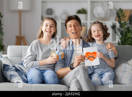 Le figlie si congratulano con il padre e gli danno una cartolina. Papà e ragazze sorridono e abbracciano. Vacanza in famiglia e insieme. Foto Stock