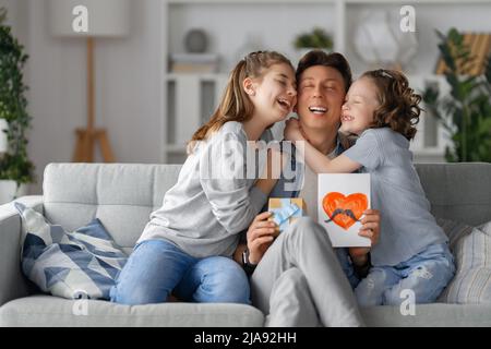 Le figlie si congratulano con il padre e gli danno una cartolina. Papà e ragazze sorridono e abbracciano. Vacanza in famiglia e insieme. Foto Stock