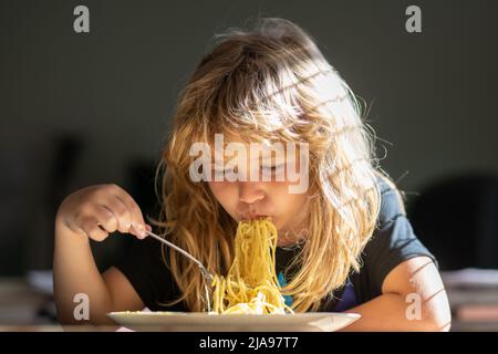 Piccolo bambino che mangia spaghetti in cucina. Bambino giovane seduto sul tavolo che mangia cibo sano con espressione divertente sul viso. Primo piano ritratto di divertente Foto Stock