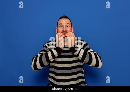 Latino uomo con le mani sulla bocca sentendosi terrore autentico, gli morde le unghie per cercare di calmare le sue paure. Isolato su sfondo blu studio. Foto Stock